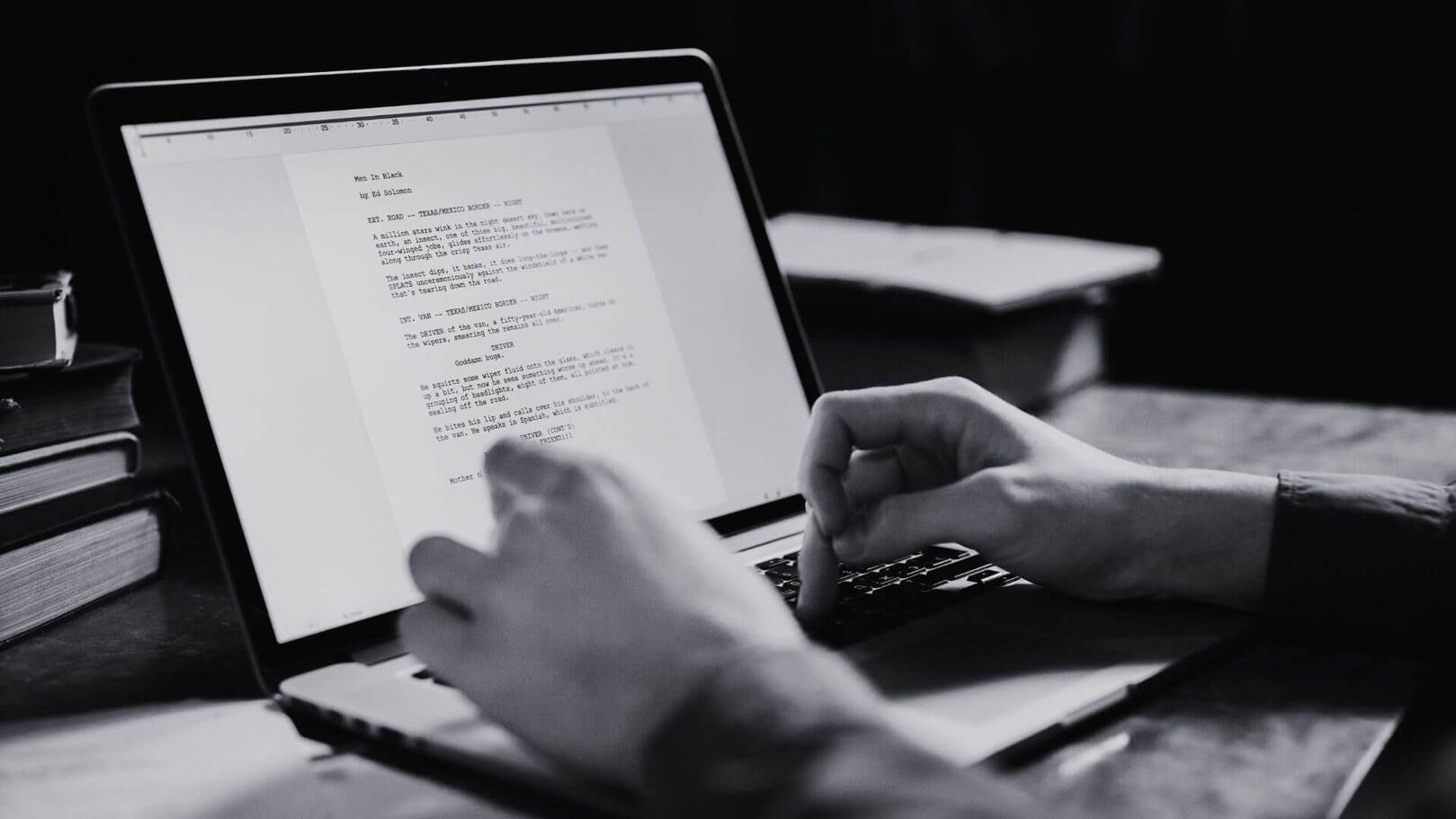 Screenwriting & Concept Development — Jessica Evelyn Rodrigues de Lima Black and white close-up of hands typing a script format screenplay on a laptop, with a glowing screen and blurred books in the background.