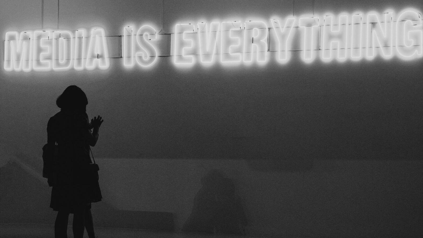 Promotion & Media Planning — Media Strategy & Content Distribution | Jessica Evelyn Rodrigues de Lima A silhouette of a person seen from behind, observing a bright glowing neon sign that reads: 'MEDIA IS EVERYTHING'.